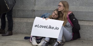 Zwei Mädchen sitzen aneinander gelehnt und halten ein Schild, auf dem „#LoveLikeJo“ steht