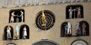 Drehfiguren in einem Uhrturm zeigen verschiedene Berufe, dazwischen sitzt ein goldener Hahn
