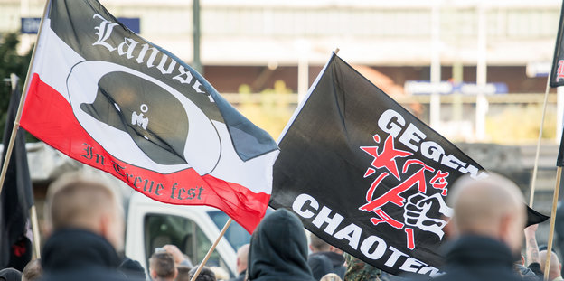 zwei Fahnen über einer Menschenmenge, eine ist schwarz-weiß-rot und hat die Aufschrift "Landser - in Treue fest", die andere ist schwarz mit der Aufschrift "Gegen Chaoten"