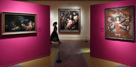 Links und rechts rosa Wände, an denen Gemälde hängen, dahinter geht eine Frau an einem dritten Gemälde vorbei