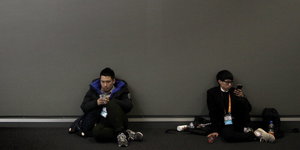 Zwei Jungen sitzen an eine graue Wand gelehnt und spielen mit ihren Smartphones