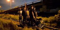 Eine Gruppe dunkel gekleidter Männer steht um einen Wagen auf Schienen, auf dem zwei in Tücher gehüllte Leichen zu sehen sind