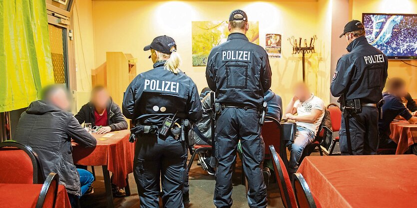 Polizisten stehen in einem Raum, an dem Männer an Tischen sitzen