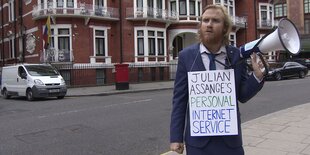 ein Mann mit Schild um den Hals und Megafon in der Hand an einer Straßenecke