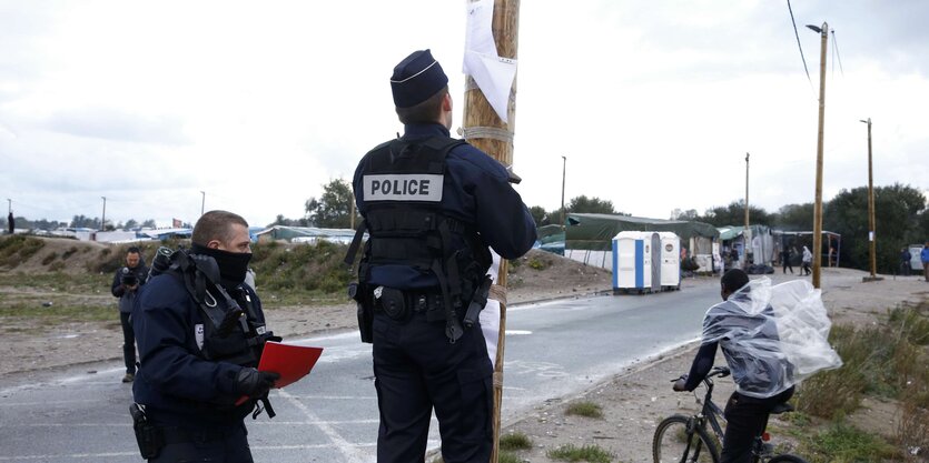 Polizisten an einem Strommast, ein Mann fährt auf einem Fahrrad vorbei, im Hintergrund Hütten