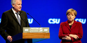 Sehofer steht an einem Rednerpult. Merkel daneben. Im Hintergrund eine blaue Wand mit dem Schriftzug CSU