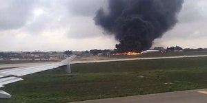 Aus einem Flugzeugfenster beim Start fotografiert und deshalb etwas unscharf: Ein Brand plus Rauchwolke auf dem Flugfeld in einiger Entfernung. Jemand löscht.