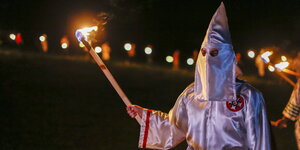 Eine Person mit spitzem weißem Hut, der auch das Gesicht verdeckt, hält eine brennende Fackel