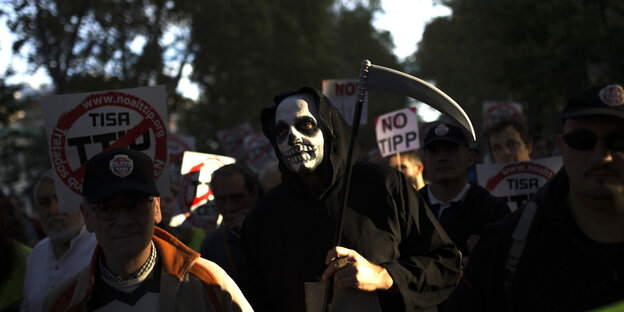 Menschen mit Anti-TTIP-Plakaten, dazwischen einer, der mit einer Totenmaske, schwarzer Kutte und Sense