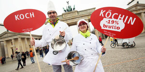 Ein Koch und eine Köchin in Arbeitskleidung halten vor dem Brandenburger Tor Schilder mit der Aufschrift „Koch“ und „Köchin -19% brutto“ in die Luft