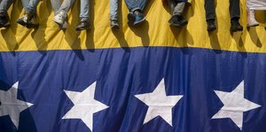 DemonstrantInnen sitzen auf einer Mauer in den Farben der Nationalflagge Venezuelas. Nur die Beine sind zu sehen