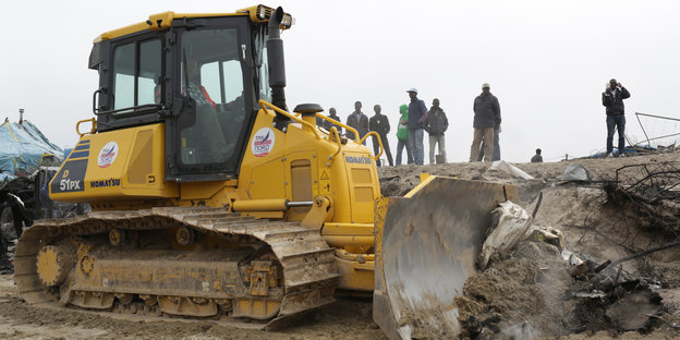 Ein gelber Bulldozer beginnt schiebt Dreck vor sich her, im Hintergrund auf einer Düne sind Flüchtlinge