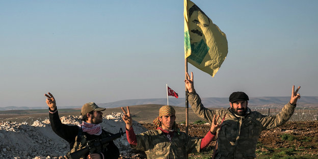 Drei YPG-Kämpfer strecken triumphierend Arme und eine Fahne in die Höhe