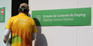 Ein Helfer im gelben Shirt geht auf eine Tür zu, daneben hängt ein Schild mit der Aufschrift: Dopingkontrolle