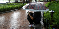 Ein Mann hockt mit einem Regenschirm im Park und spielt auf seinem Smartphone