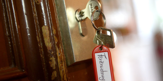 Eine Tür, von der der Lack abplatzt. Im Türschloss steckt ein Schlüssel mit einem roten Anhänger, auf dem „Ferienwohnung“ steht