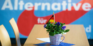 Auf einem Tisch steht ein Korb mit violetten Blumen, darin ein Deutschlandfähnchen, dahinter an der Wand das AfD-Logo