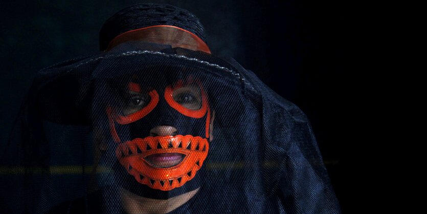 Ein Mensch trägt eine rote Lucha Libre Maske, darüber einen Hut mit einem dunklen Schleier