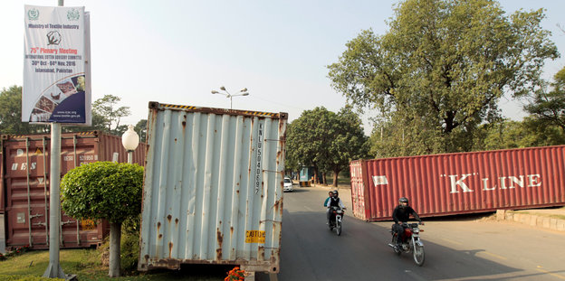 Ein graue und einer rötlicher Container liegen schräg auf einer Straße, dazwischen fahren Menschen auf Motorrädern
