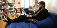 Ein junger Mann sitzt mit ausgestreckten Beinen auf einer Couch und schreibt auf einem Laptop