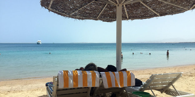 Auf Liegestühlen liegen zwei Menschen unter einem Sonnenschirm mit Blick auf das blaue Meer