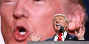 Donald Trump steht redend hinter einem Pult, hinter ihm ist eine Großaufnahme seines Gesichts