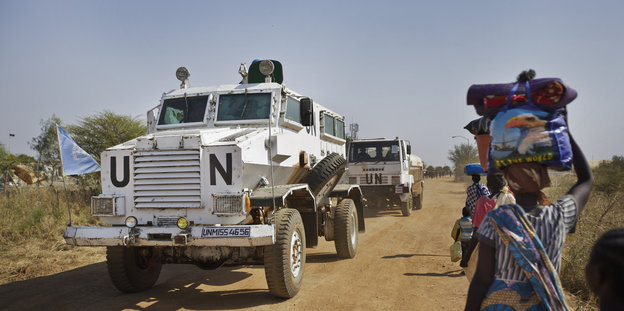 Ein weißes Fahrzeug fährt über eine Sandstraße. Auf dem steht UN. Daneben tragen Frauen Sachen auf dem Kopf.