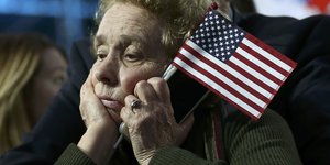 Eine enttäuscht schauende ältere Dame hält eine Amerikaflagge und ein Smartphone in der linken Hand. Sie stützt den Kopf auf die rechte Hand.