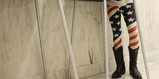 Eine Person mit Leggins mit dem Motiv der amerikanischen Flagge darauf und Cowboystiefeln an, steht hinter einer Wahlkabine Eine Person mit Leggins mit dem Motiv der amerikanischen Flagge darauf und Cowboystiefeln an, steht hinter einer Wahlkabine