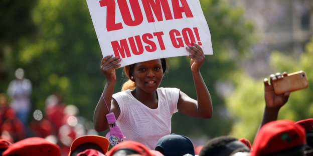 Auf einer Demo hält eine Frau ein Plakat hoch. Auf diesem steht „Zuma must go“
