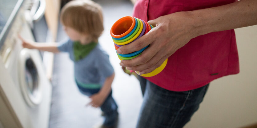 Ein Kind steht auf der Boden, seine Mutter hält ein Spielzeug in der Hand