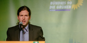 Ein Mann in Anzug und mit langen, zurückgebundenen Haaren spricht vor einem Podium, hinter ihm ein grünes Plakat