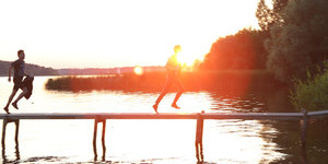 Zwei Männer rennen über einen Steg, der über Wasser führt, dahinter ein Sonnenuntergang