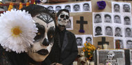 Zwei Menschen haben ihre Gesichter zu Totenschädeln geschminkt, hinter ihnen an der einer Wand hängen Fotografien