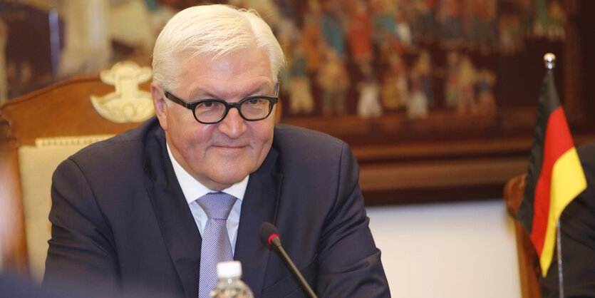 Ein Mann mit grauen Haaren und Brille sitzt vor einem Mikrofon, rechts im Bild ist die deutsche Nationalflagge zu sehen