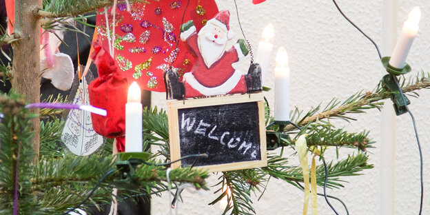 An einem Weihnachtsbaum hängt eine Tafel mit der Aufschrift „Welcome“, auf der ein hölzerner Weihnachtsmann sitzt