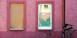 Die Hauswand eines rosa angestrichenen Hauses, vor einem der zwei Fenster ist eine Holzplatte angebracht