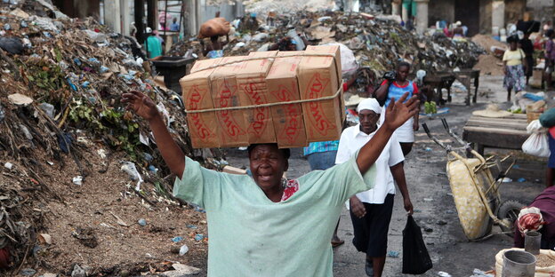 Eine Frau trägt ein großes Paket auf ihrem Kopf, um sie herum Berge von Müll und Trümmern
