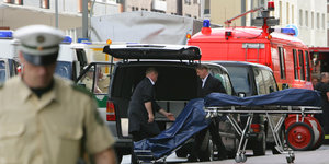 Ein Polizist verschwommen im Vordergrund, im Hintergrund mehrere Rettungswagen und ein Leichenwagen, in den gerade zwei bedeckte Leichen gelegt werden