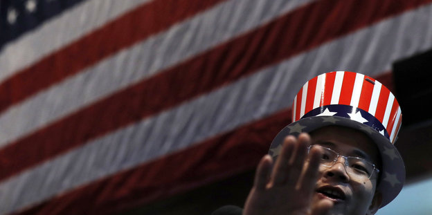 Ein Mann mit Hut in Farben der US-Flagge winkt. Hinter ihm hängt auch noch eine US-Flagge