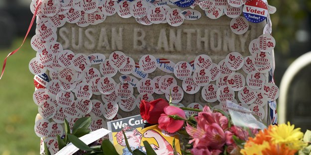 Auf einem Grabstein kleben viele Aufkleber „I voted today“, davor liegen Blumen