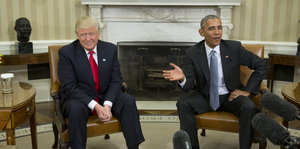 Zwei Männer in Anzügen sitzen auf Stühlen nebeneinander, im Hintergrund ein Kamin