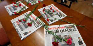 Auf einem Holztisch liegen verschiedene Ausgaben einer Zeitung, auf ihnen mehrere rote Nelken
