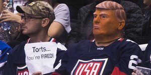 Zwei Menschen sitzen in Eishockey-Trikots auf der Tribüne, einer hat eine Maske auf, die aussieht, wie das Gesicht von Donald Trump und hält ein Blatt Papier in der Hand