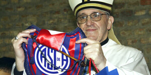 Ein Mann mit Brille und in Kardinalsgewand und -mütze hält einen Wimpel eines Fußballvereins hoch