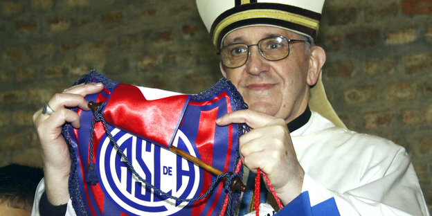 Ein Mann mit Brille und in Kardinalsgewand und -mütze hält einen Wimpel eines Fußballvereins hoch
