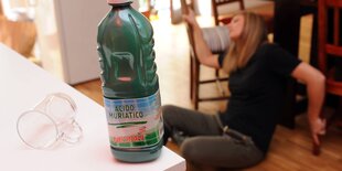 Im Vordergrund auf der Tischplatte: eine Flasche Salzsäure, im Hintergrund eine auf dem Boden liegende Frau