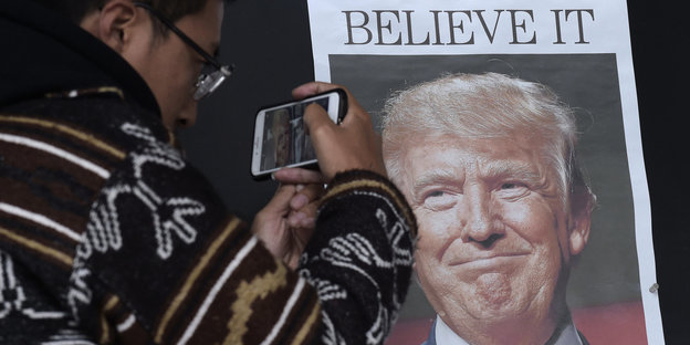 Ein mann fotografiert eine Zeitungsseite mit Trumpbild und der Zeile „Believe it“