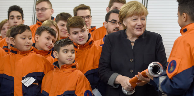 Merkel vor mehreren Kindern in Feuerwehr-Klamotten. Sie hält einen Löschschlauch