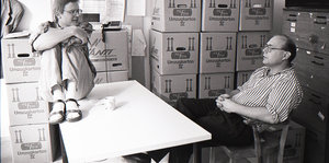 Ein Raum voller Umzugskartons. Arno Widmann sitzt am Tisch, Karl-Heinz Ruch kauert auf der Tischplatte